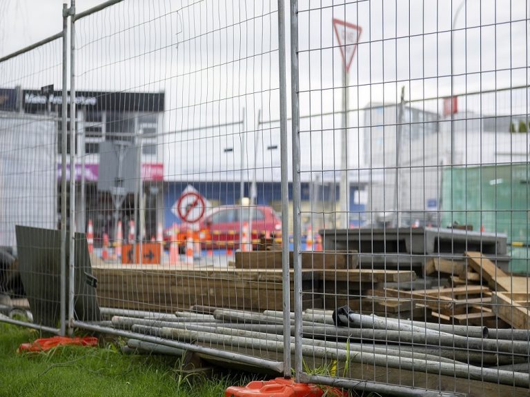 Construction materials protected by metal fence. Out-of-focus traffic cones and cars in the distance. Roadworks in Auckland.